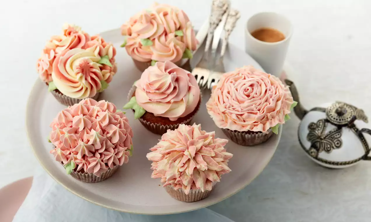 Blumen-Cupcakes mit pastellfarbenem, zu Blüten gespritztem Frosting, frischen Himbeeren und kleinen Minzblättern auf weißem Porzellanteller