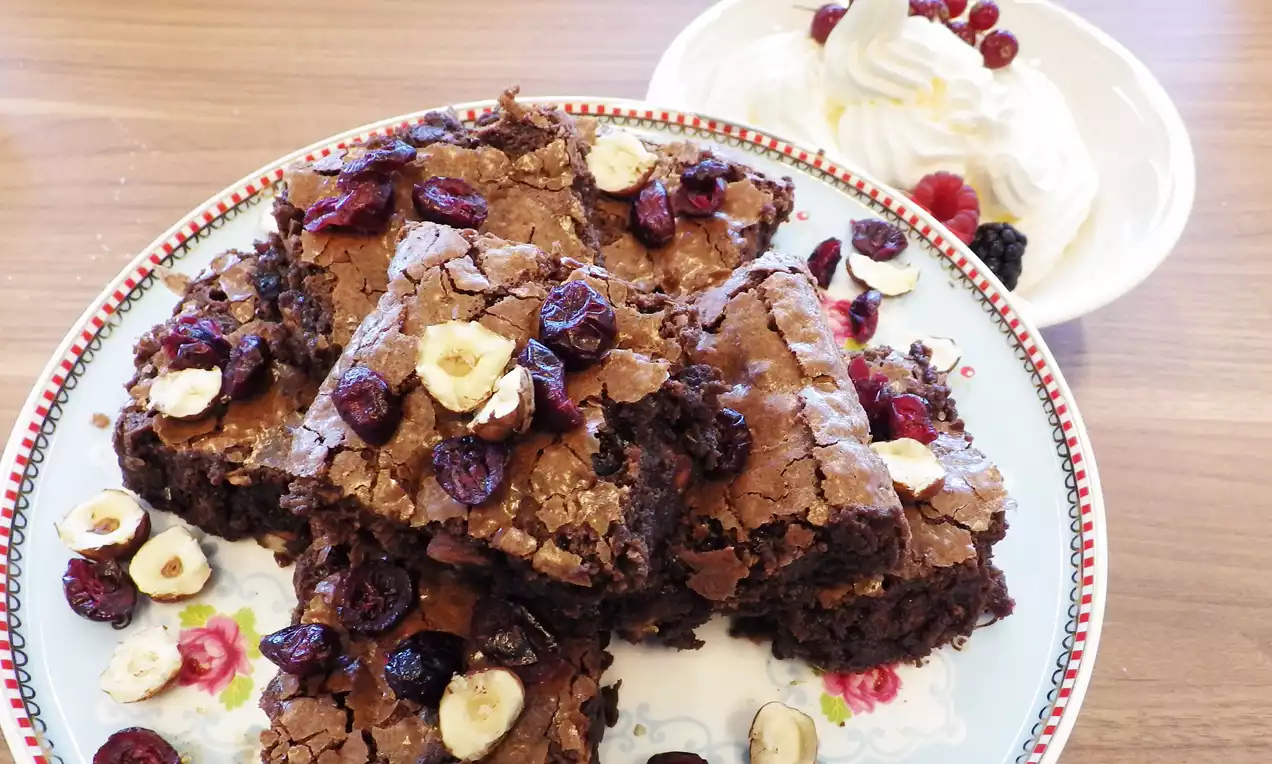 Glanzende, donkerbruine brownies met een krokante bovenkant, bestrooid met hazelnoten en rode cranberries op een sierlijk bloemmotiefbord