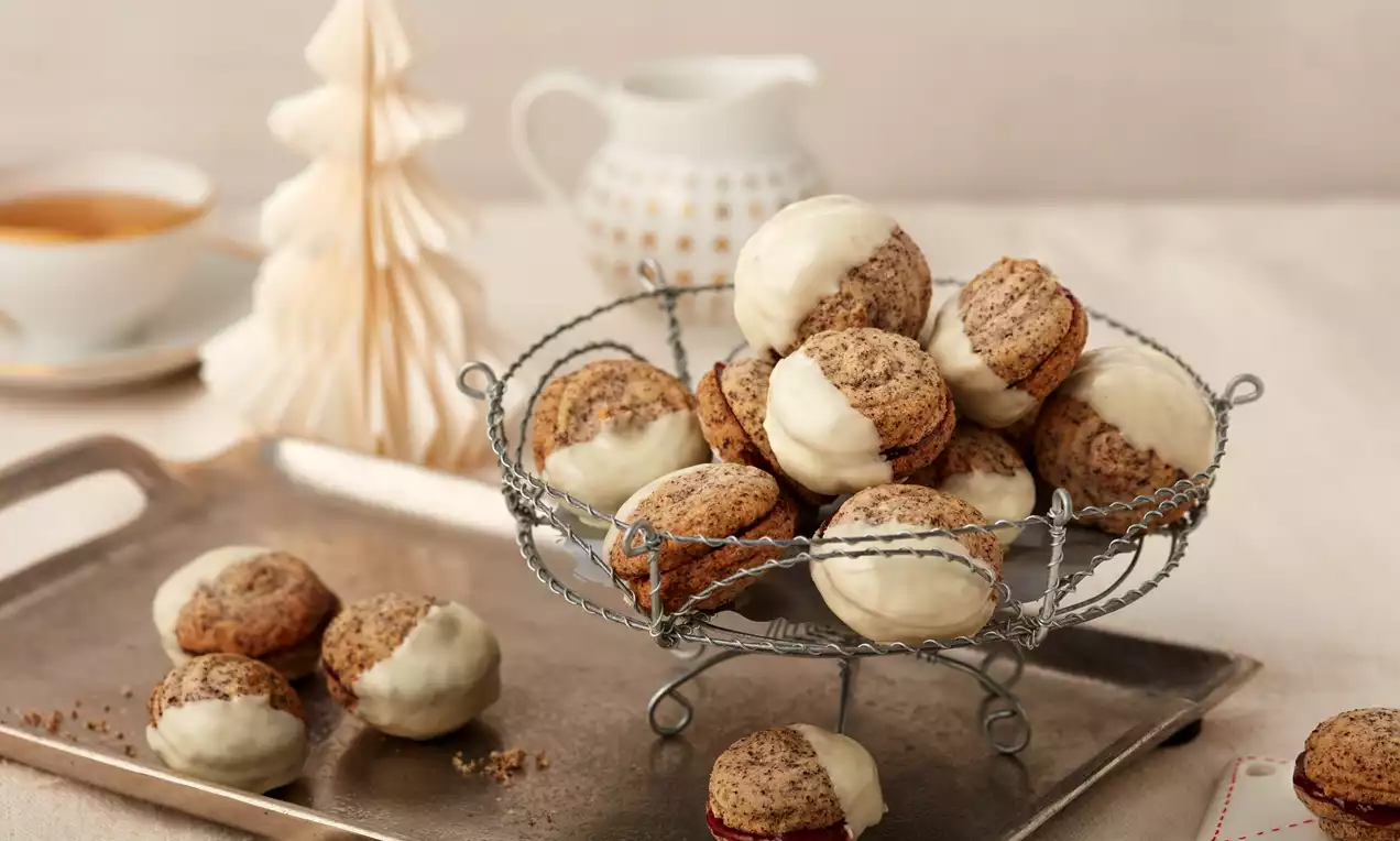 Gefüllte Mohnplätzchen mit hellem Boden und dunkler Mohnschicht, teils mit weißer Glasur, in silbernem Korb auf Tablett mit winterlicher Deko