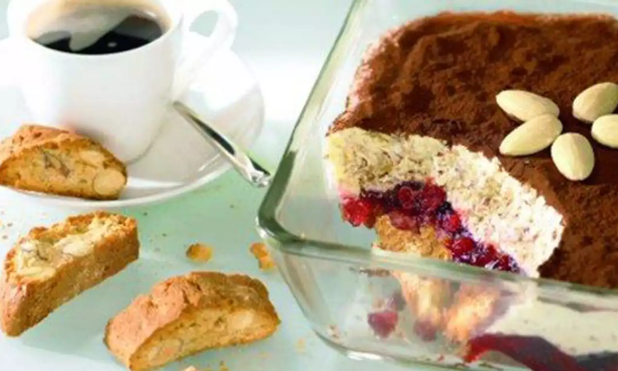 Tiramisu ve skleněné nádobě s vrstvou kakaového prášku, červeným ovocem a mandlovou dekorací, vedle biscotti a šálku kávy