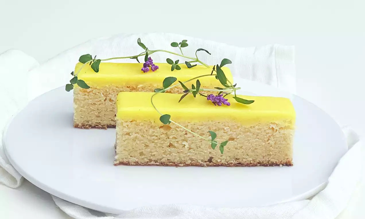 To lysegule citronkagestykker med glat marcipanoverflade og dekorative grønne kviste med lilla blomster på en hvid tallerken