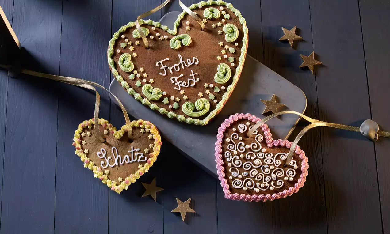 Drei braune Lebkuchenherzen mit bunter Zuckerdekoration und Schrift, mit goldener Schleife auf dunklem Holz und kleinen Deko-Sternen