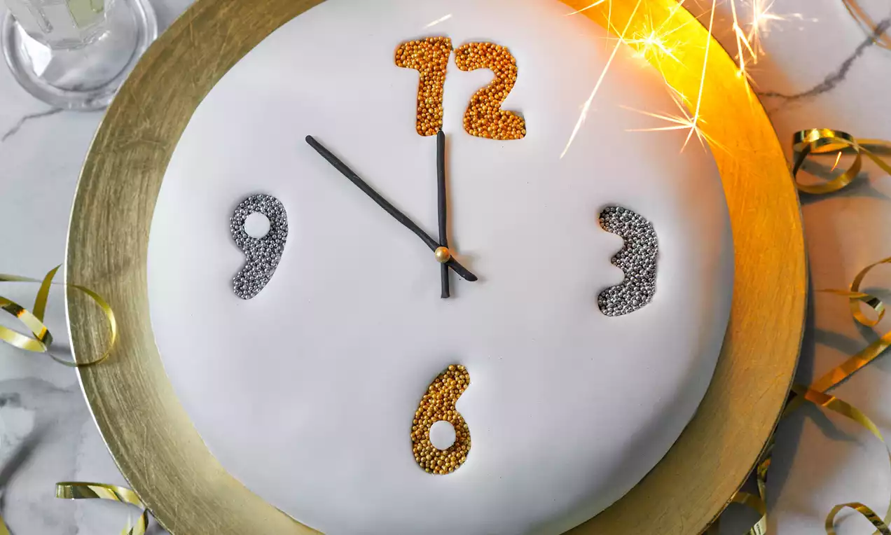 Okrúhla torta s hladkým bielym poťahom, číslicami zo zlatých a strieborných cukrových perličiek, čiernymi ručičkami a zlatým tanierom