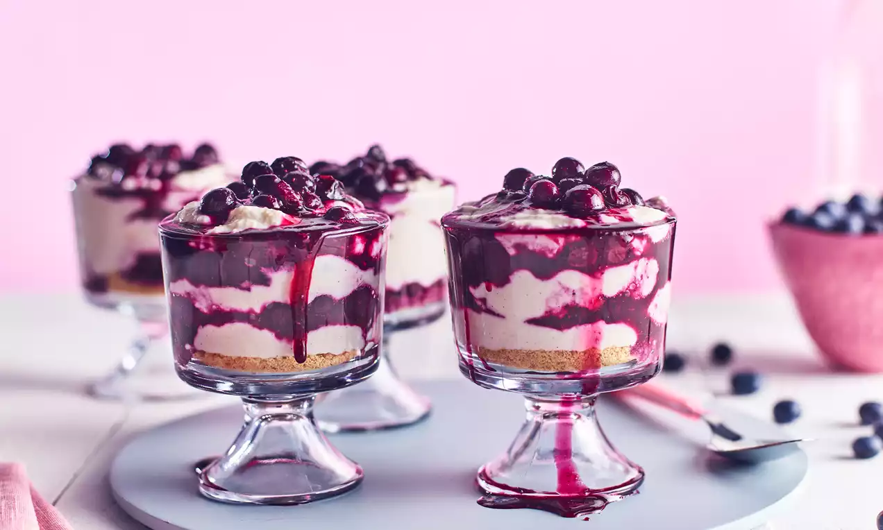 Layered blueberry cheesecake in a glass with cream and biscuit base, vibrant purple blueberry topping and syrup drips on a pastel backdrop