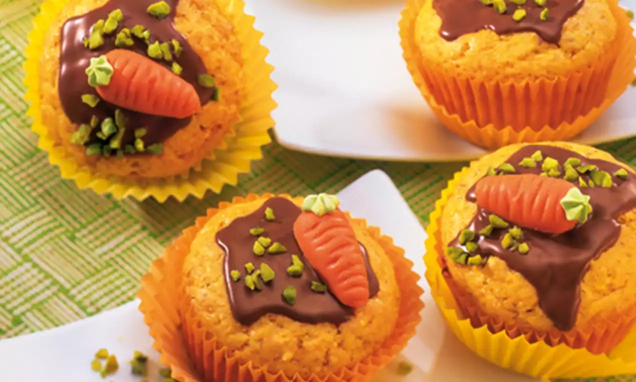 Muffins met warme oranje tinten in gele papieren vormpjes, gegarneerd met chocolade, een marsepeinen wortel en groene pistachestukjes