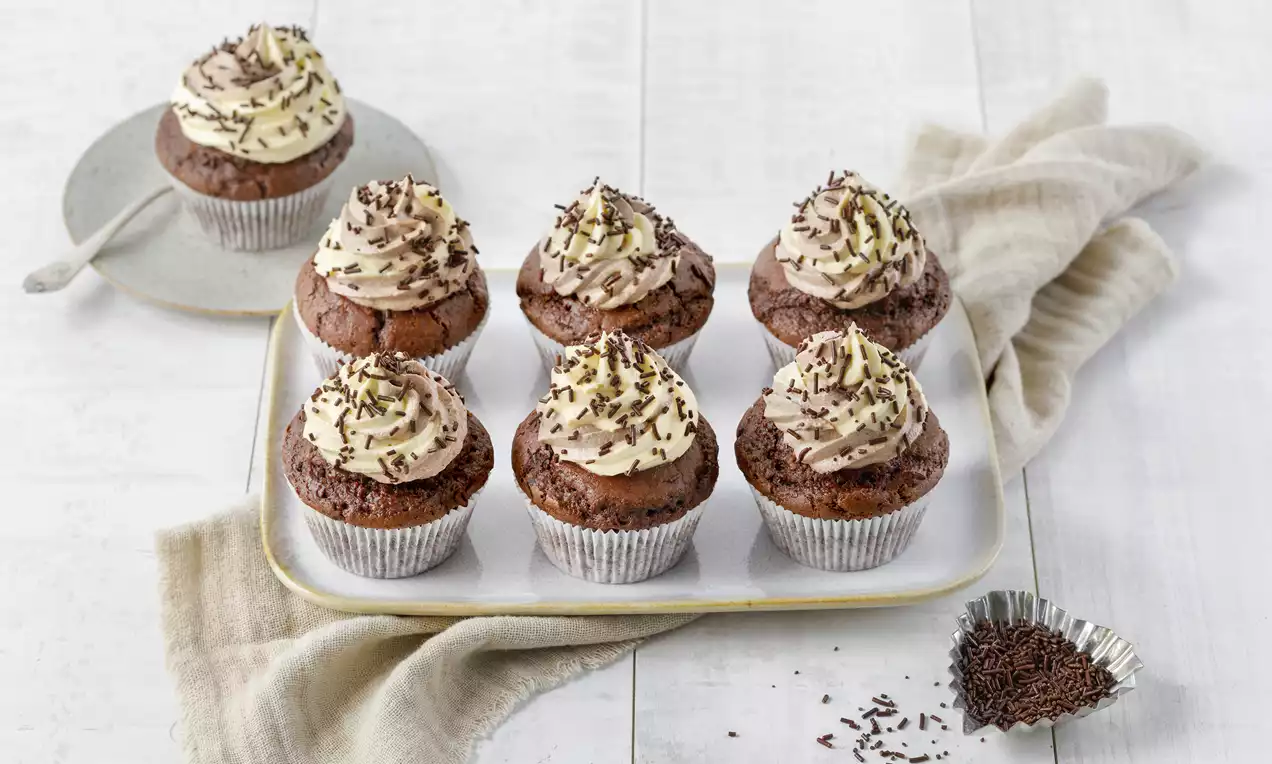 Vegane Schoko-Cupcakes mit cremigem Topping, helle und dunkle Schokostreusel auf einem weißen Tablett, umgeben von beigem Stoff