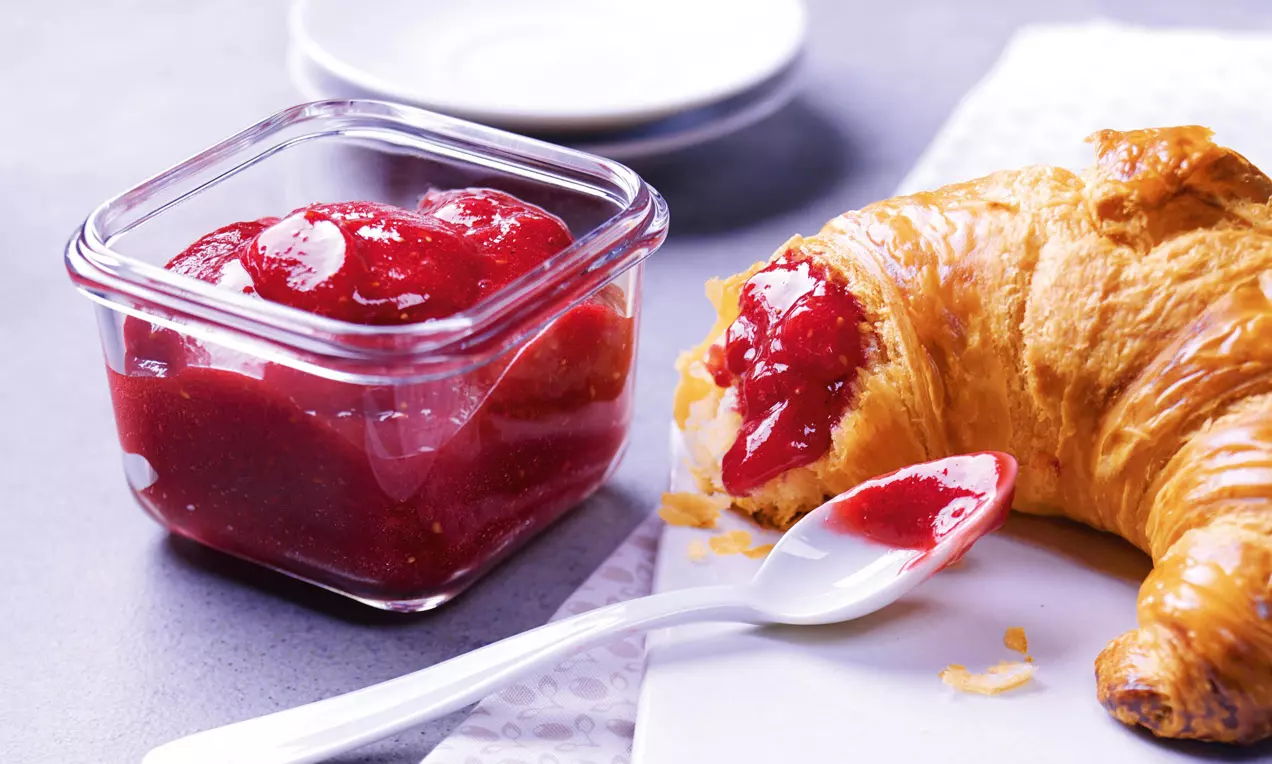 Jasně červený džem s hladkou texturou ve skleněné nádobě, vedle dozlatova upečeného croissantu a bílé plastové lžičky na světlém povrchu