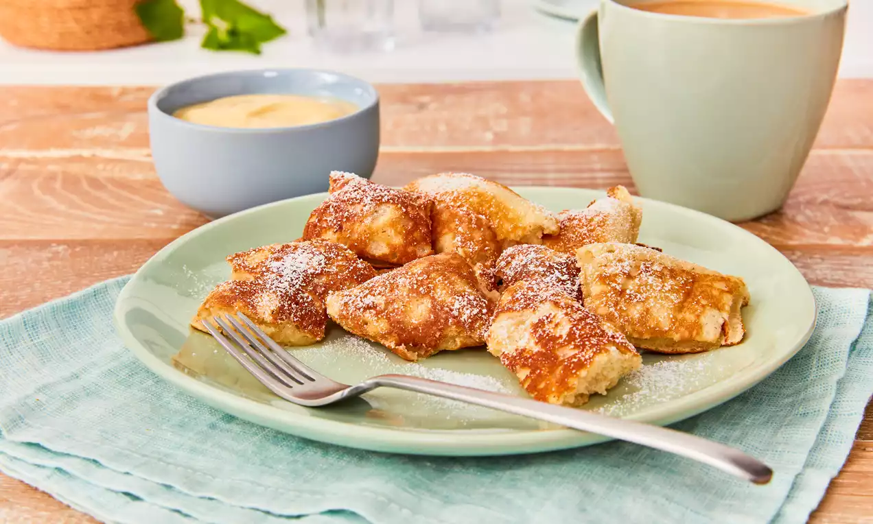 Komadići zlatnosmeđeg, mekanog drobljenca posuti šećerom u prahu na svijetlozelenom tanjuru uz vilicu, umak i šalicu kave