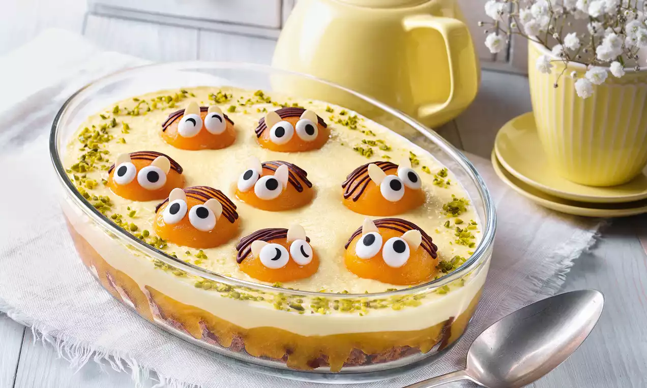 Ovale Glasform mit gelblichem Pudding, darauf Aprikosenhälften mit Zuckeraugen und Schokostreifen, umrandet von gehackten Pistazien