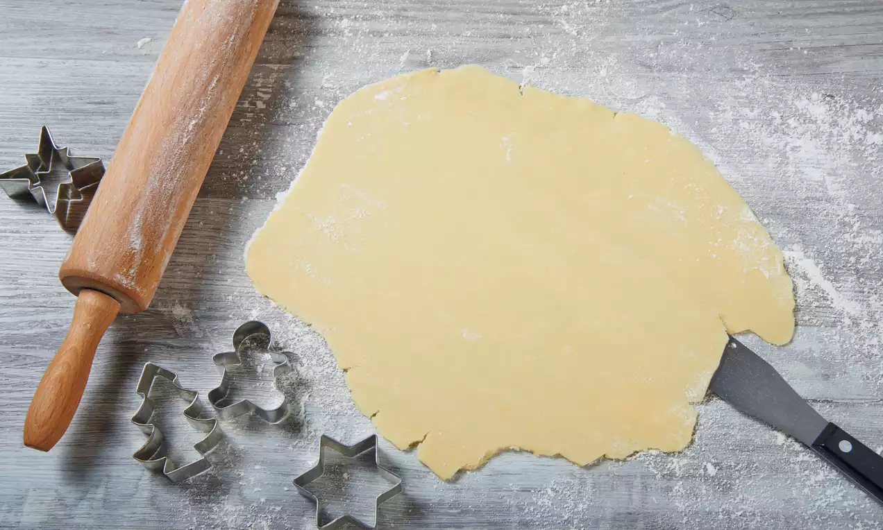 Vaaleankeltainen taikinapinta kaulittuna ohueksi harmaalla puupöydällä, vieressä metallisia piparkakkumuotteja ja puinen kaulin