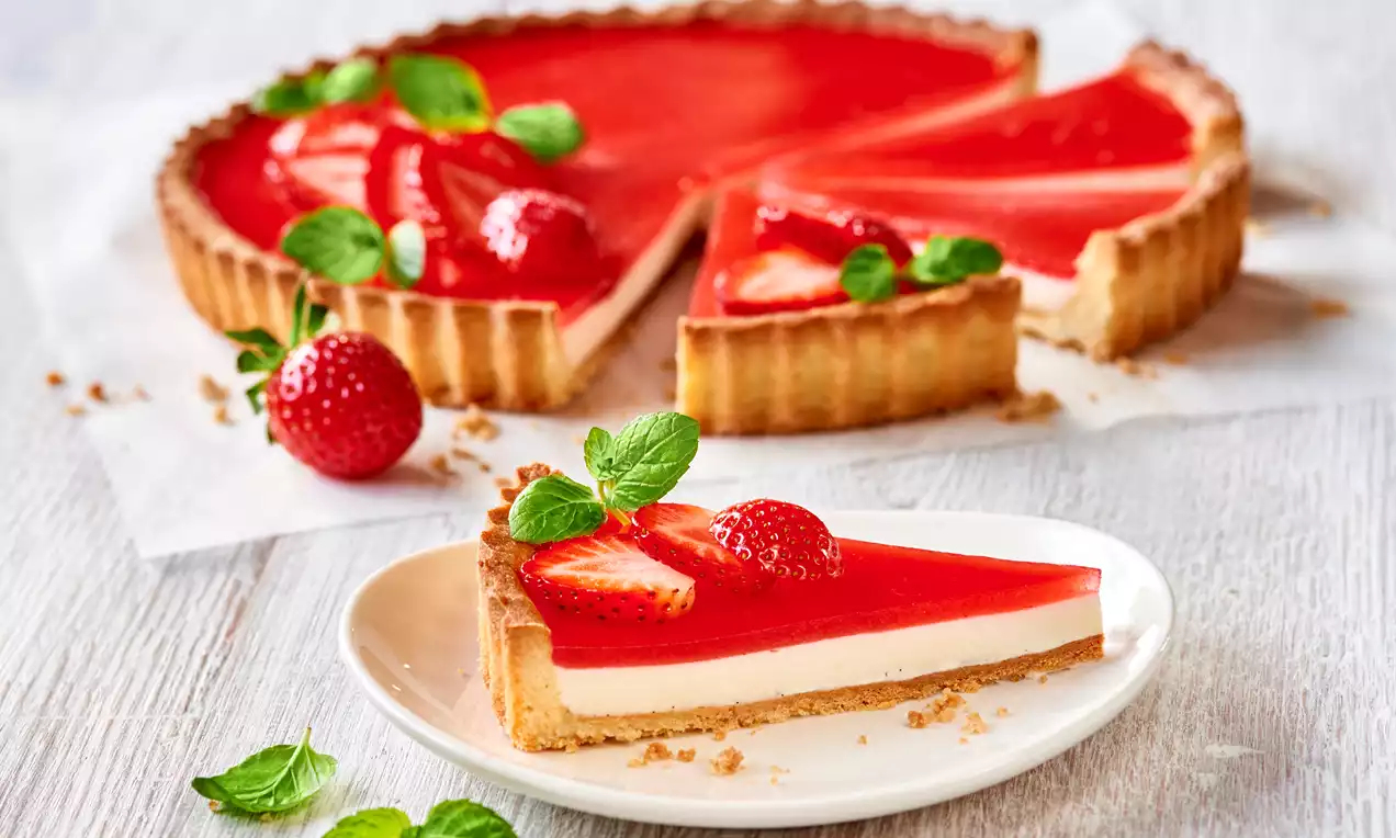 Tart s hrskavim zlatnosmeđim rubovima, glatkim slojem bijele vanilije panna cotte i sjajnom crvenom jagodinom glazurom, ukrašen svježim jagodama i listićima mente na bijelom tanjuru
