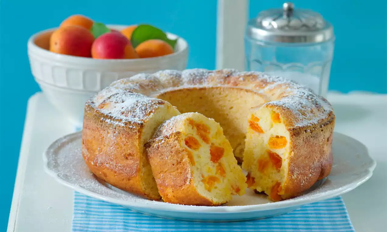 Tvarohový věnec ve zlatavém odstínu, posypaný moučkovým cukrem, s viditelnými kousky jasně oranžových meruněk na bílém talíři