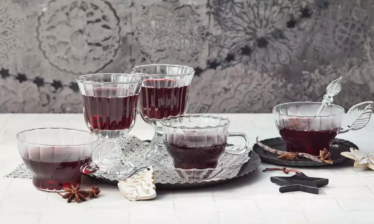 Teplé červené varené víno v čírych sklenených pohároch a šálkach, ozdobené klinčekmi, škoricou a hviezdičkami na bielom stole