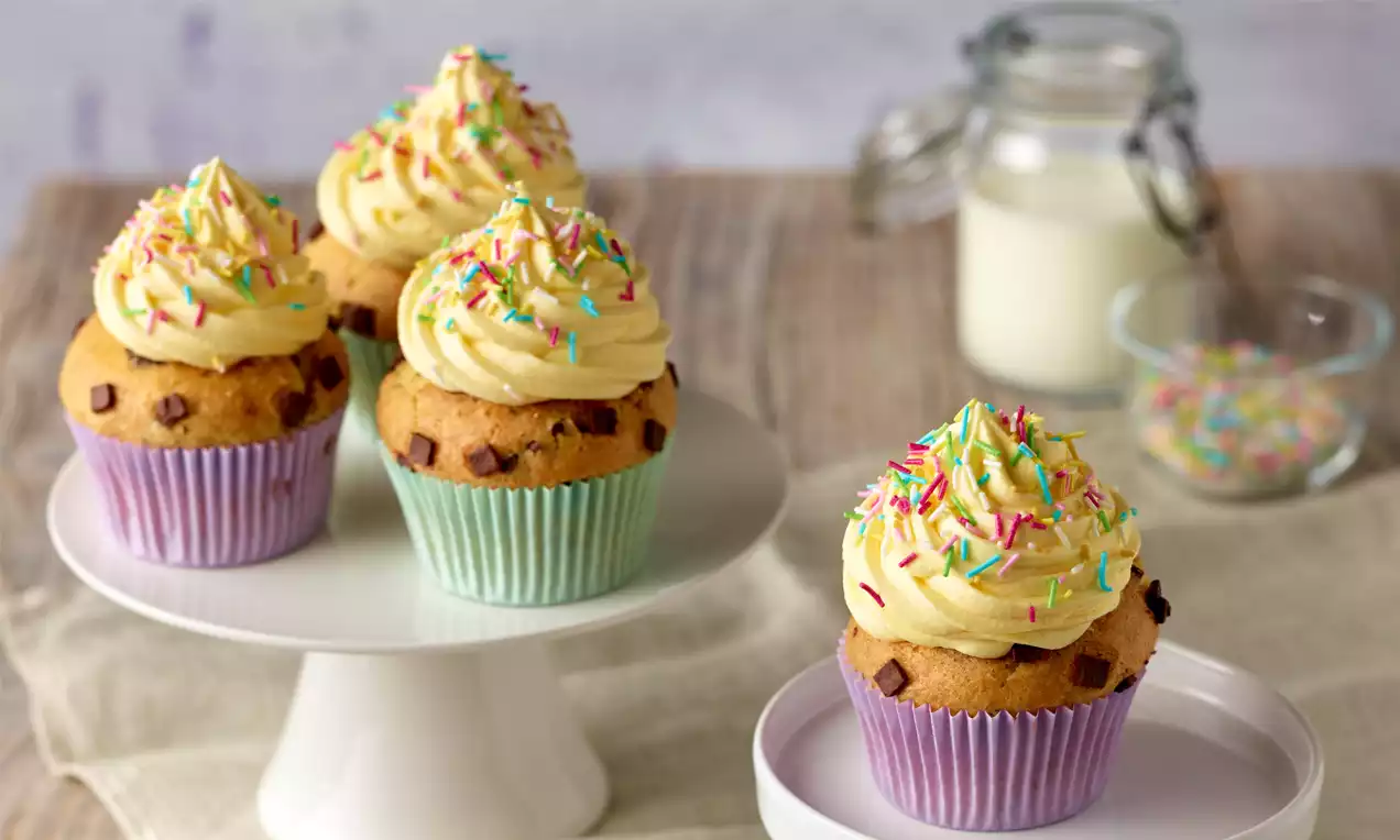 Vanillefarbene Cupcakes mit gelbem Paradiescreme-Topping und bunten Streuseln in pastellfarbenen Förmchen auf Etageren angerichtet