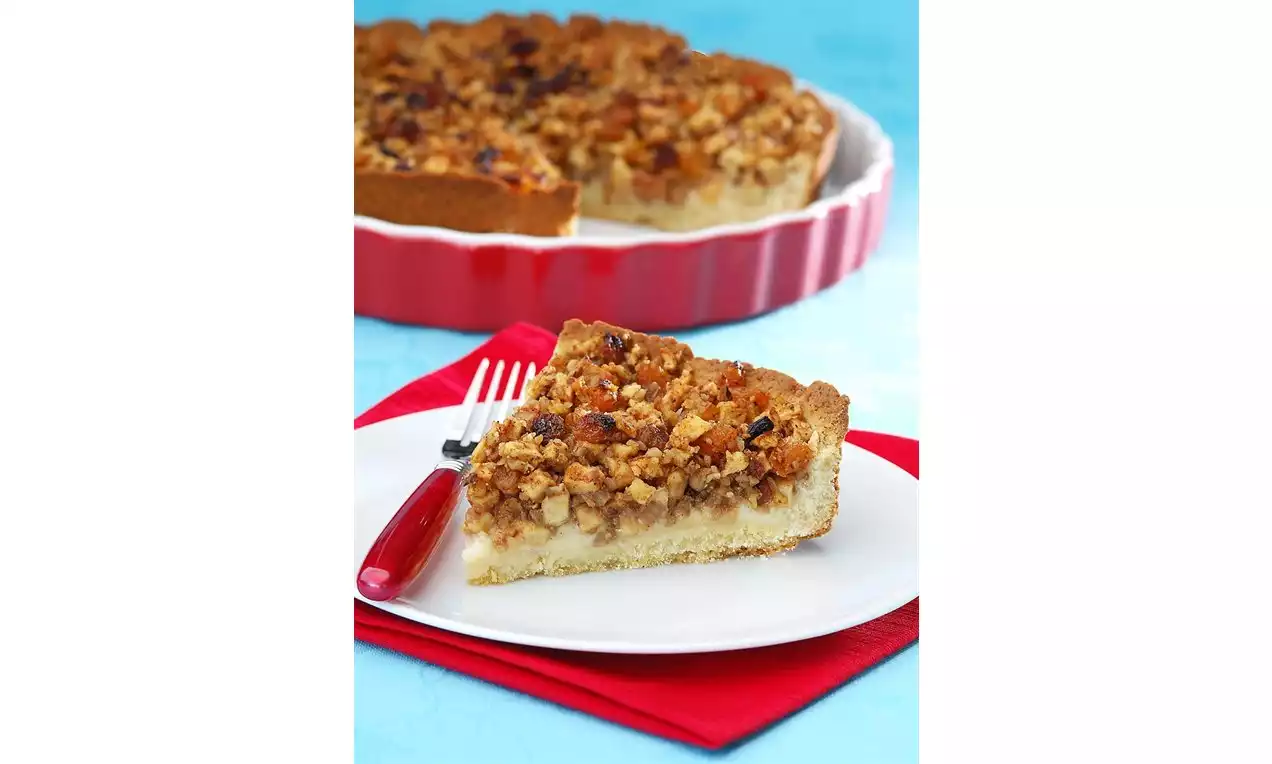 Kıtır ceviz kırıntılarıyla kaplı, altın rengi tatlı dolgulu, pürüzsüz tart hamurlu dilim beyaz tabakta ve kırmızı bir çatalla sunulmuş