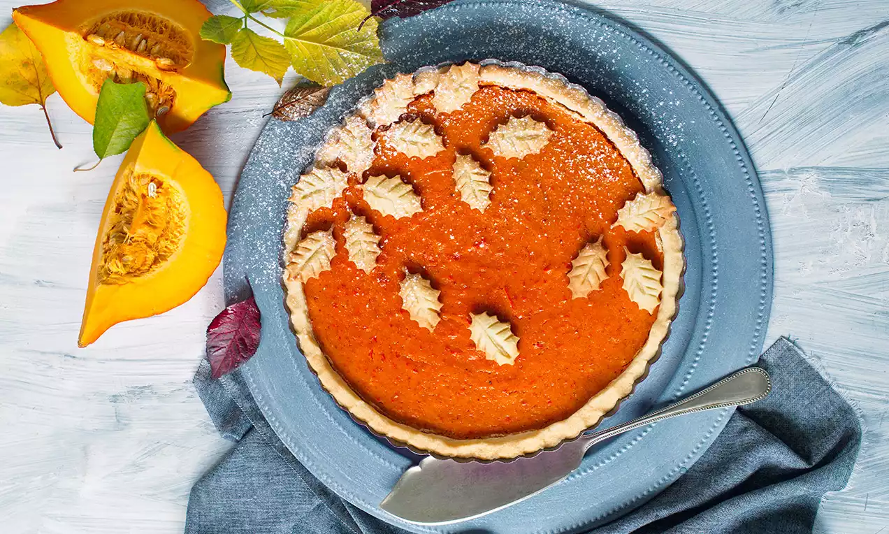 Dýňový koláč s jasně oranžovou náplní a ozdobami z krémové šlehané hmoty na světlé křehké kůrce, na modrém talíři s nožem a podzimní dekorací