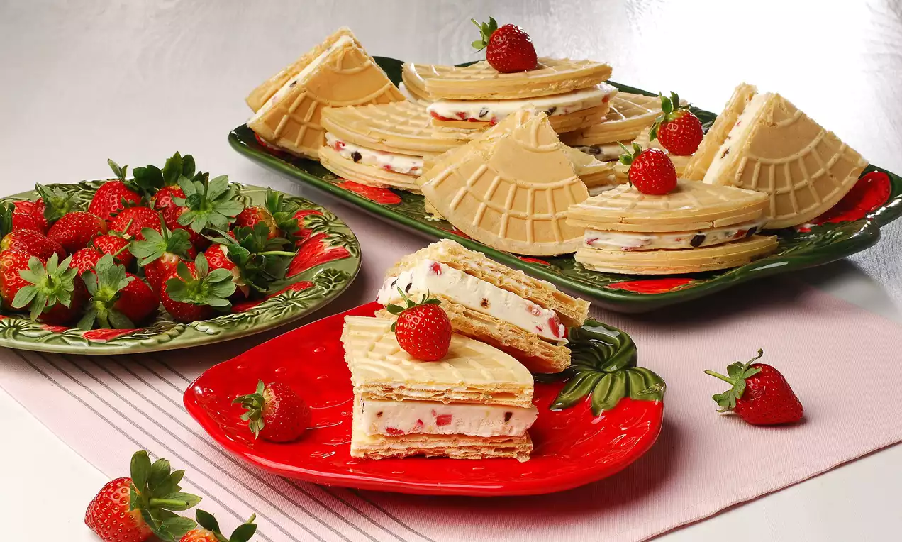 Kare dilimli kağıt helvalar, içleri vanilyalı puding ve çilek parçalarıyla dolu, yanındaki taze çileklerle renkli tabaklarda sunulmuş