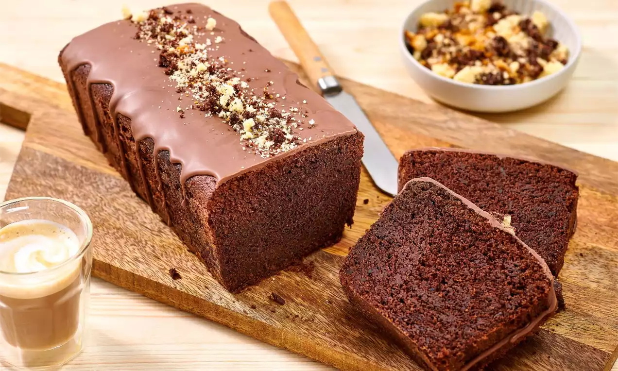 Čokoládový chlebíček s hladkou hnedou polevou, posypaný drobenkou, nakrájaný na plátky vedľa hrnčeka kávy na drevenom podnose