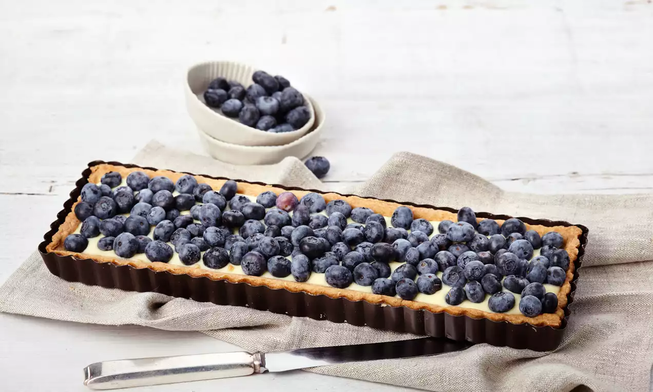 Rechteckige Blaubeer-Tarte mit goldbraunem Rand und hellem Puddingbelag, dicht bedeckt mit frischen Blaubeeren auf einem Leinentuch drapiert