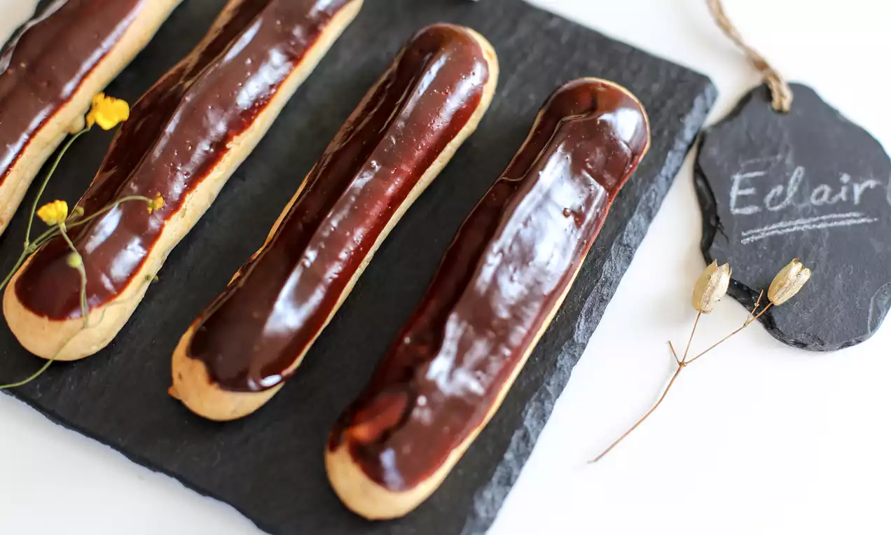 Lesklé čokoládové eclairs podlhovastého tvaru s jemne zlatistým cestom na tmavej bridlicovej doske ozdobenej žltými kvietkami a tabuľkou