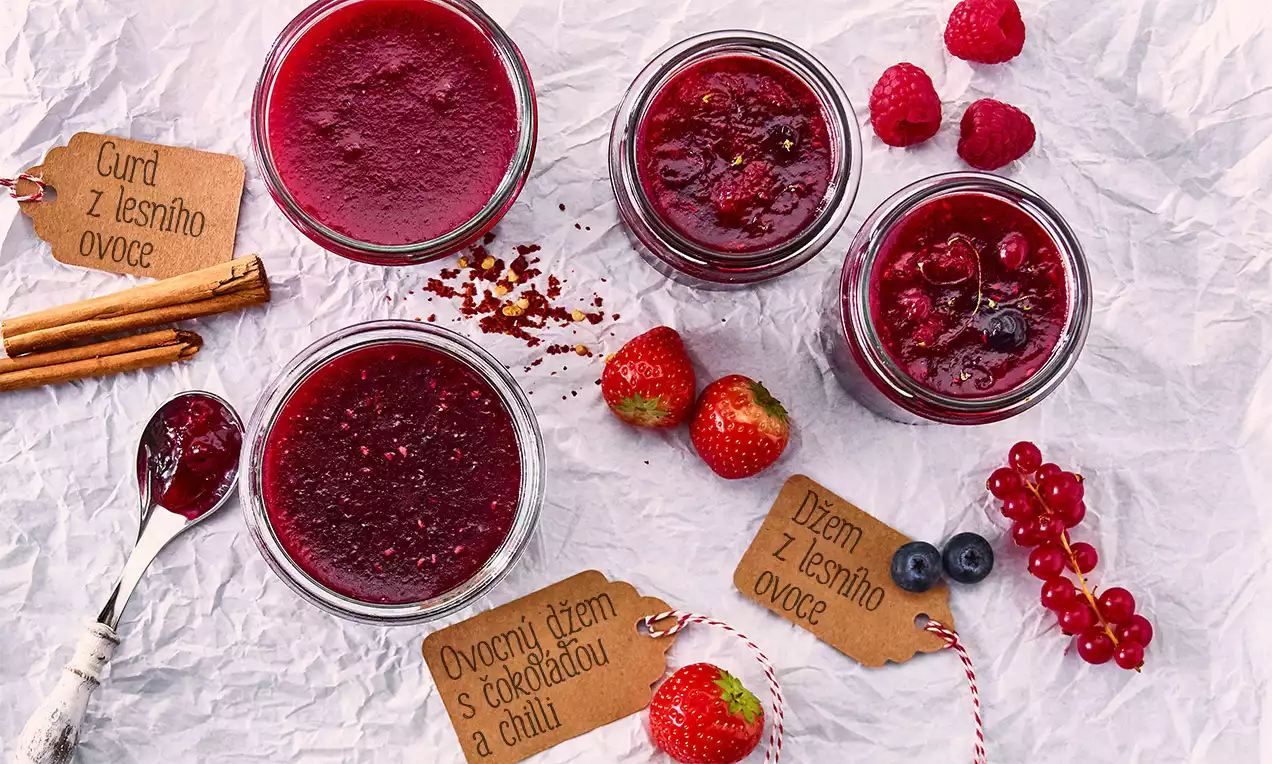 Skleničky plné tmavě červeného džemu, obklopené čerstvým lesním ovocem, skořicí a papírovými cedulkami na zmačkaném světlém podkladu