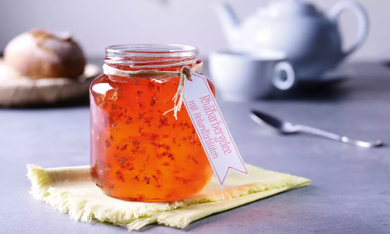 Glas mit orange-rotem Rhabarbergelee und verstreuten Holunderblüten auf gelbem Stofftuch, im Hintergrund Teekanne und Gebäck