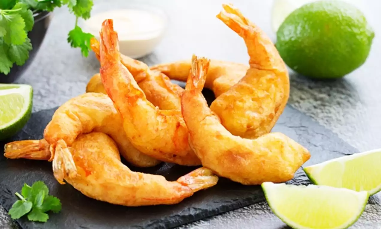 Golden crispy prawns curved with tails up, arranged on a dark slate with vibrant lime wedges, creamy dip and fresh cilantro