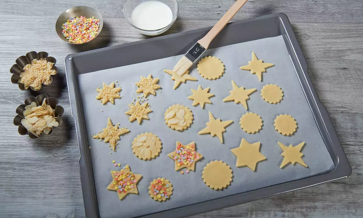 Ausstechplätzchen in Stern- und Kreisformen aus hellgelbem Teig auf Backpapier, teils bestreut mit bunten Streuseln und gehobelten Mandeln