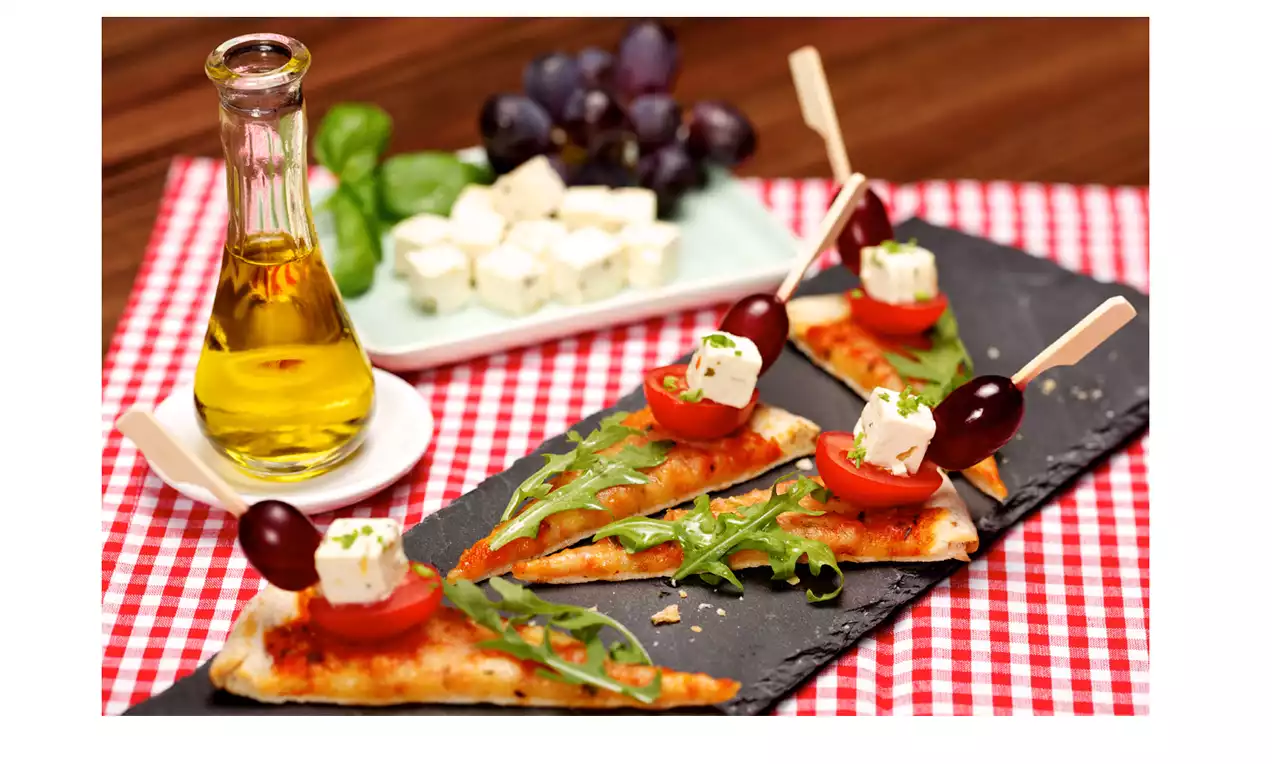 Dunne pizzapunten met rode tomatensaus, rucola, cherrytomaat, feta en olijven op zwarte plaat, naast olijfolie en feta op geruit kleed