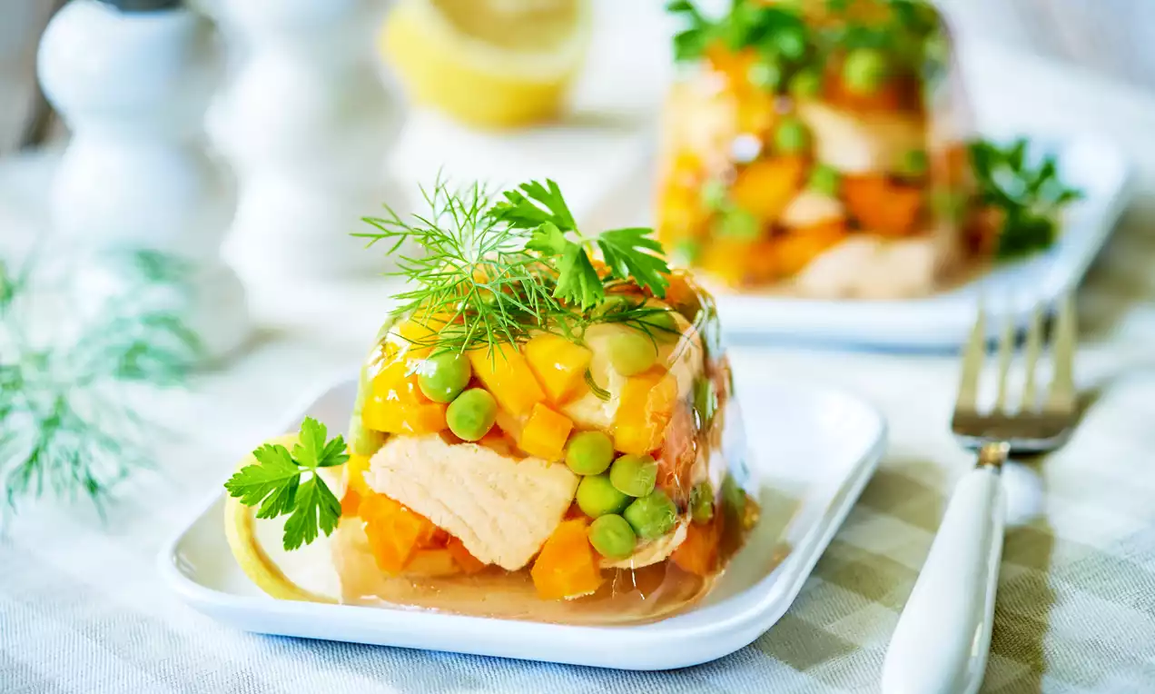 Kolorowa galaretka z łososiem, zielonym groszkiem i marchewką, udekorowana gałązkami koperku i natką pietruszki na białym talerzu