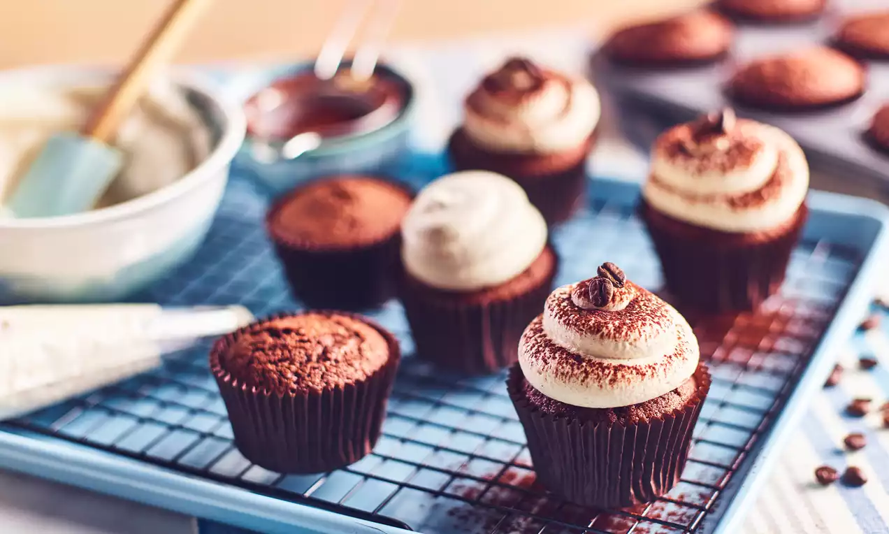Chocolate cupcakes in brown wrappers topped with smooth cream swirls, cocoa dusting, and coffee beans arranged on a blue wire rack