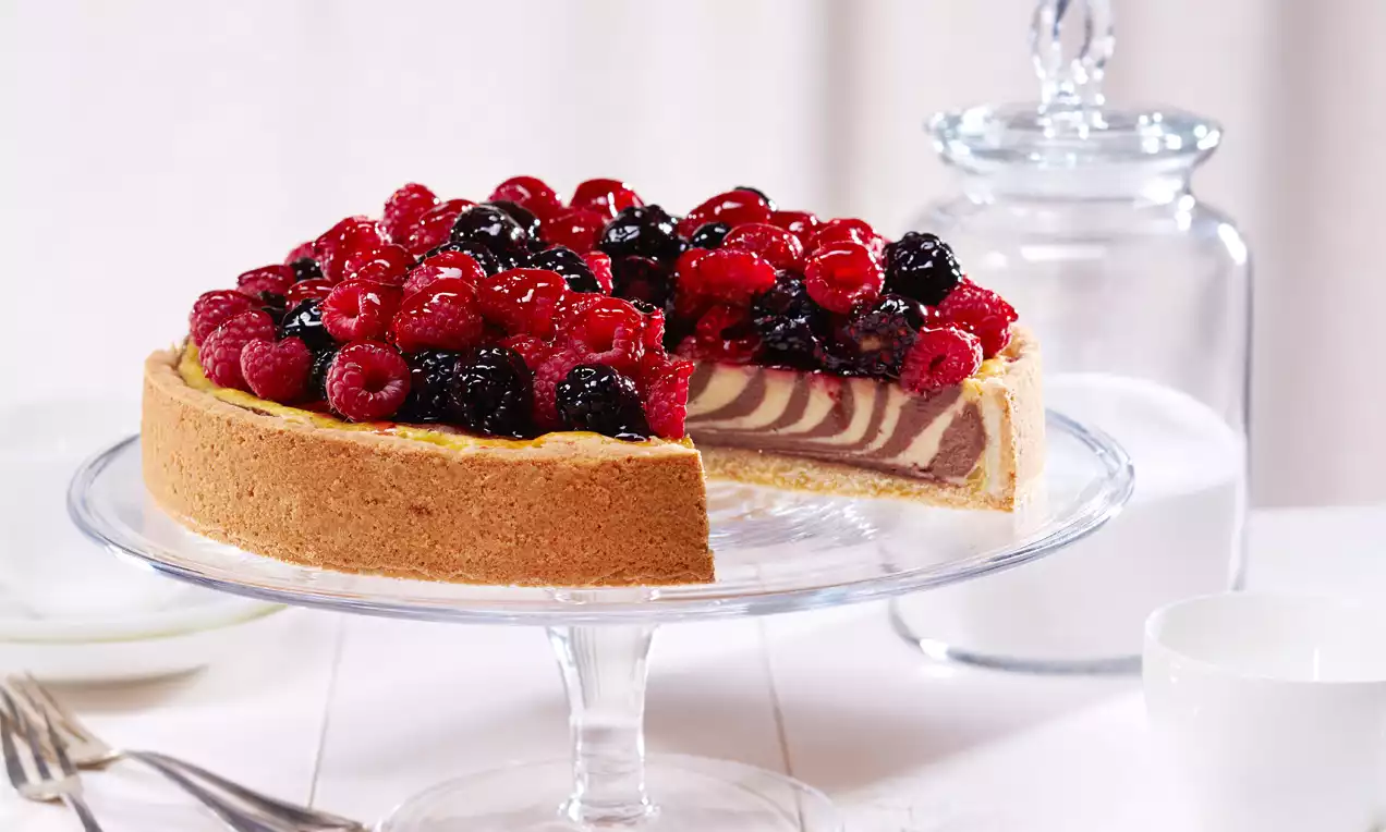 Zebra-Cheesecake mit marmorierter Füllung aus heller und dunkler Creme, bedeckt von glänzenden roten und schwarzen Beeren auf Glasplatte