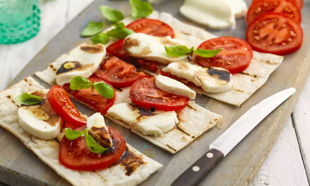 Thin flatbread squares layered with charred mozzarella slices, juicy red tomato, basil leaves, and balsamic drizzles on a wooden board