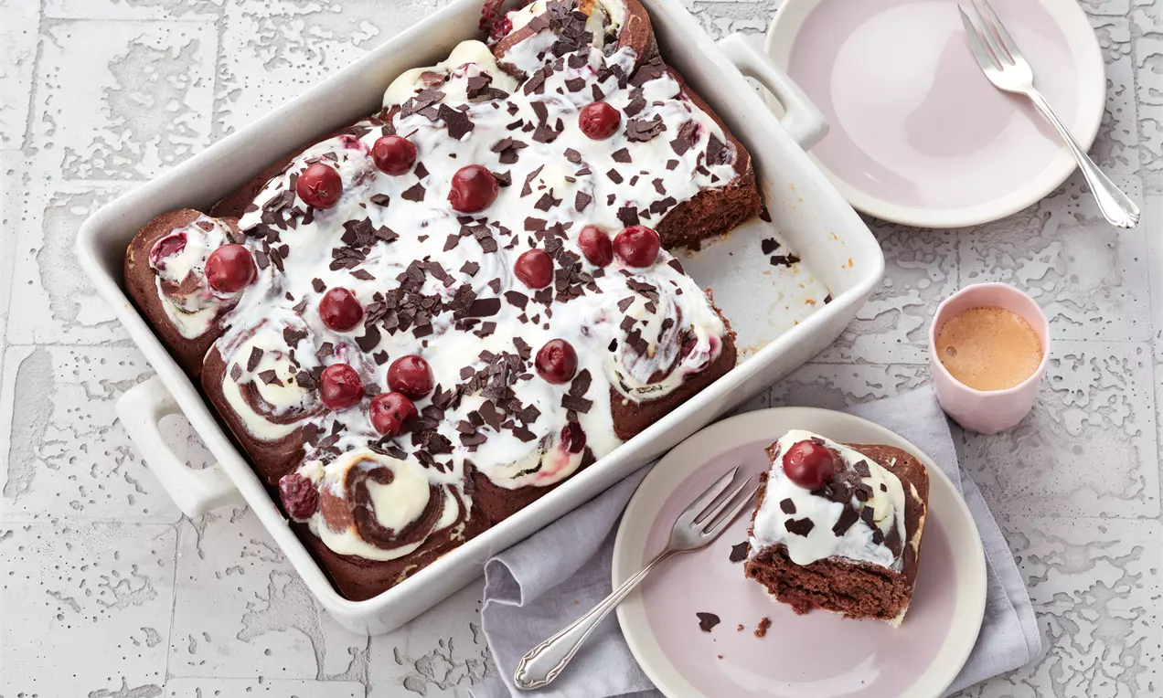 Schokoladige Schnecken in einer weißen Backform, bedeckt mit Sahne, Kirschen und dunklen Schokosplittern, dazu Teller mit Servierstück