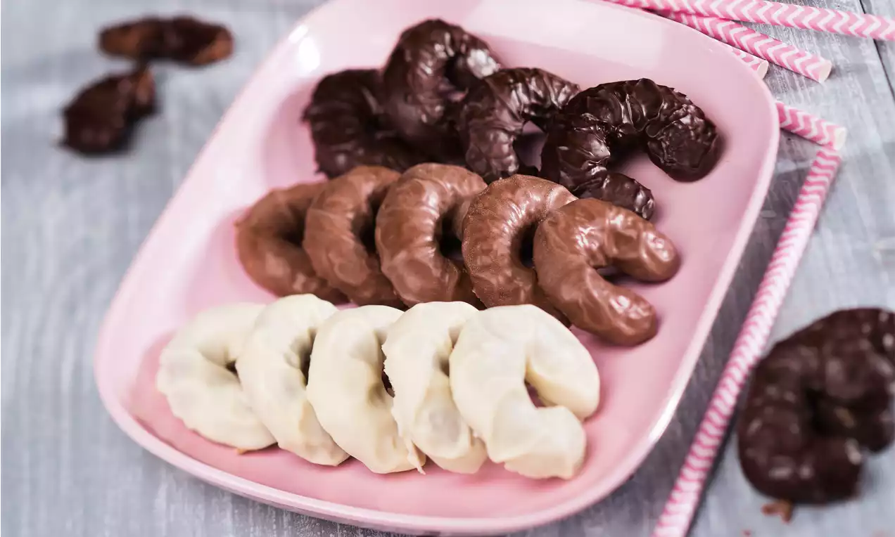 Drei Reihen Schokoladenkipferln in dunkler, heller und weißer Schokolade, dicht aneinandergereiht auf einem rosa Teller mit gestreiften Strohhalmen