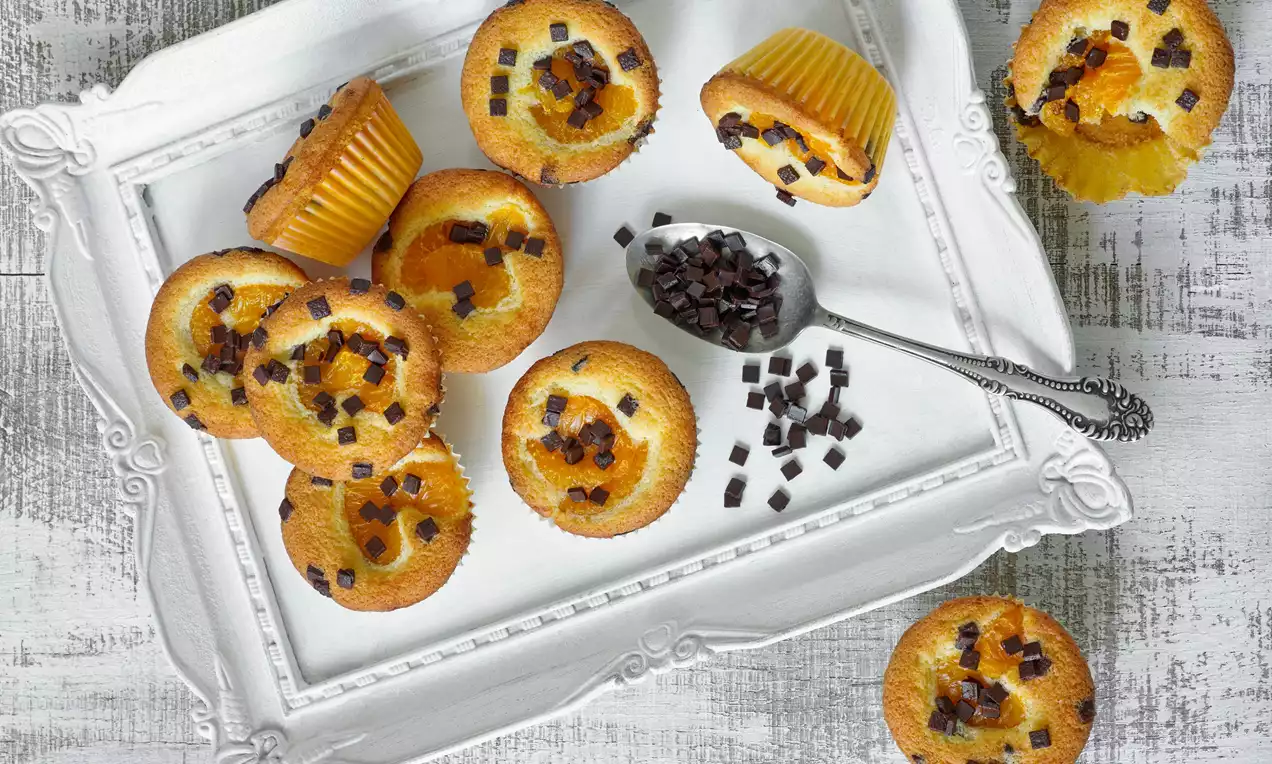 Zlatisté mandarínkové muffiny s nepravidelne rozhodenými kúskami tmavej čokolády na bielom ozdobnom podnose vedľa čajovej lyžice