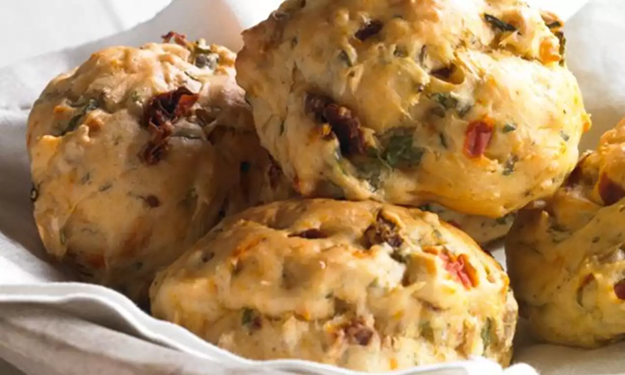 Petits pains dorés au fromage parsemés de tomates séchées et de roquette, disposés dans un panier en tissu blanc