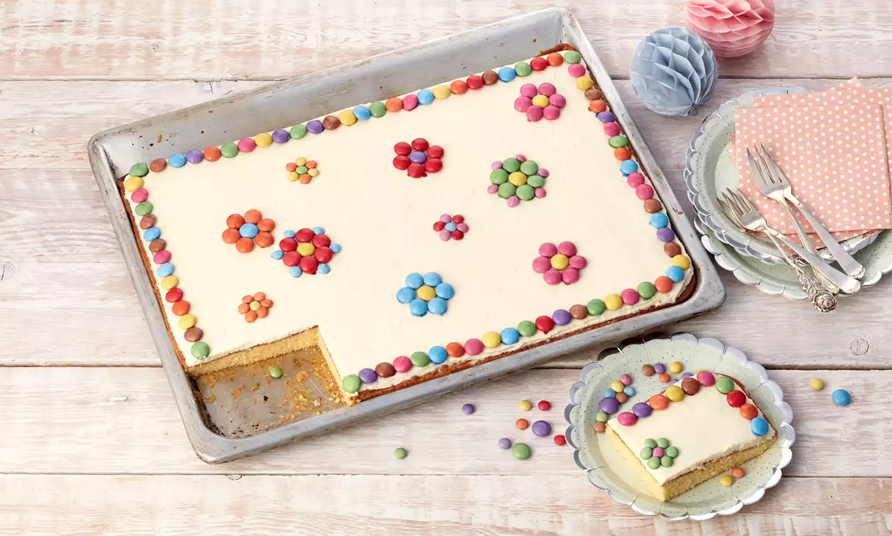 Blechkuchen mit heller Glasur, bunt verzierten Blumen aus Schokolinsen, angeschnitten und angerichtet auf einem Blumendesign-Teller