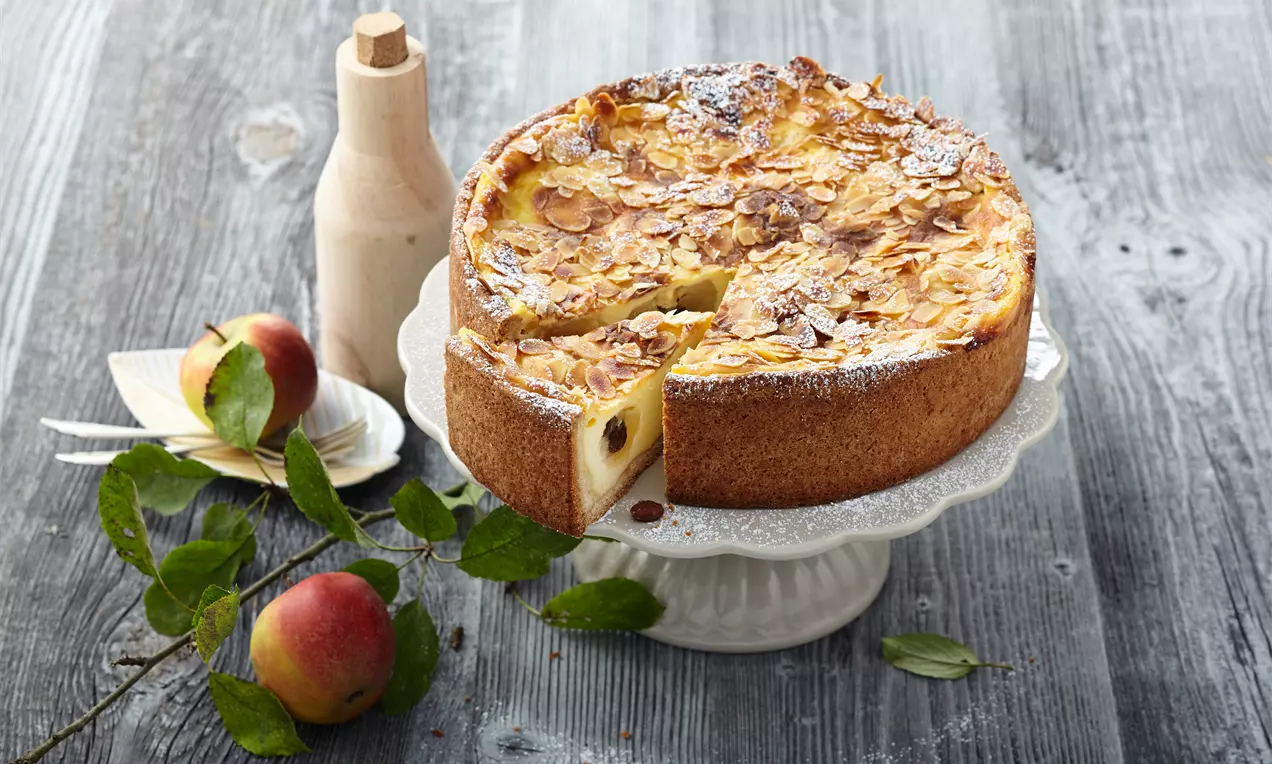 Bratapfelkuchen mit goldbrauner Kruste, cremiger Puddingfüllung, karamellisierten Apfelscheiben und dunklen Rosinen auf weißem Teller