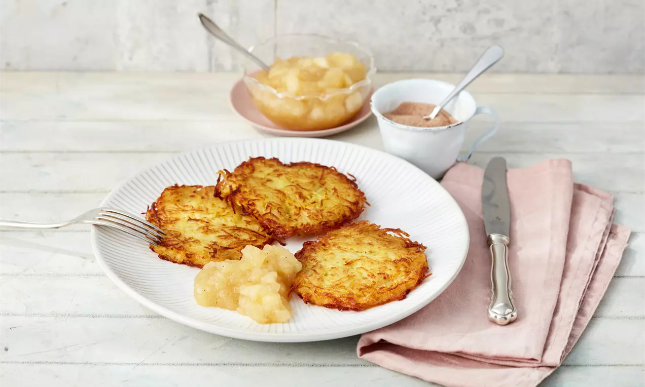 Kartoffelpuffer aurii și crocanți, așezați pe o farfurie albă alături de piure de mere, un cuțit argintiu și șervețel roz pal