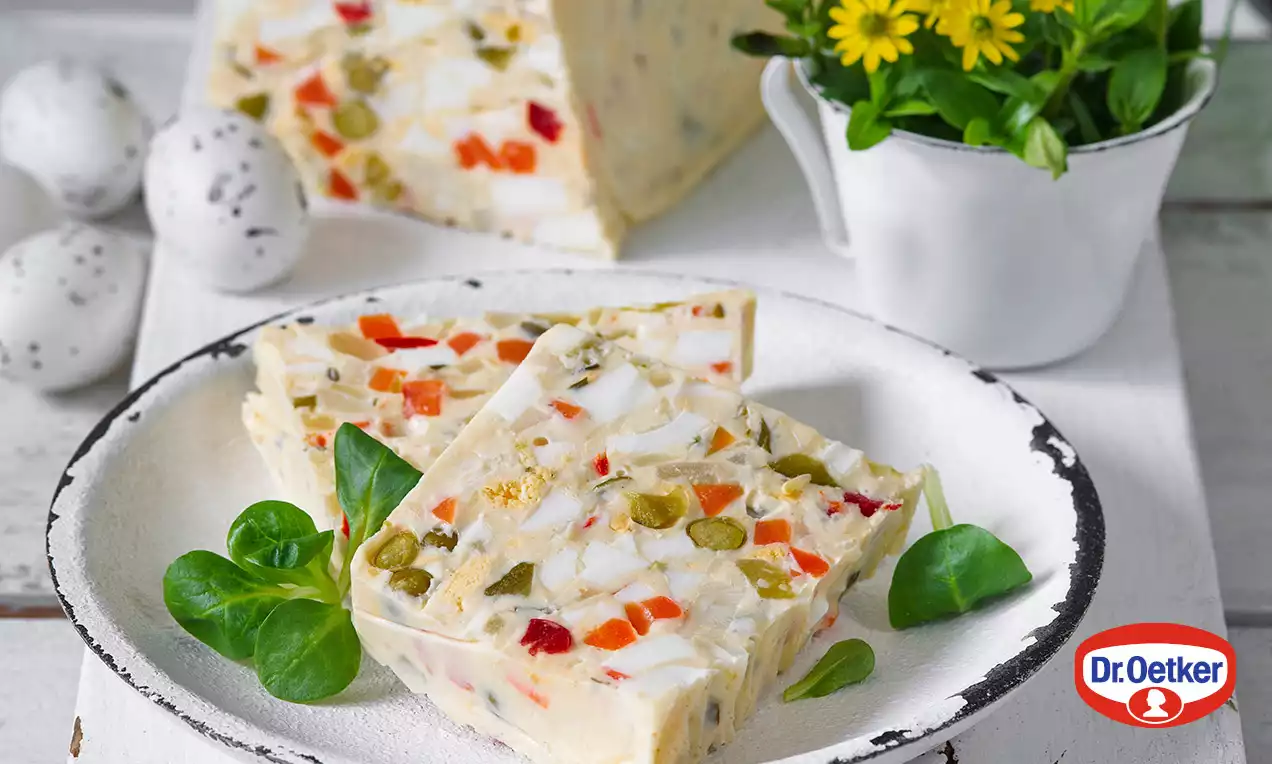 Jemne krémová tlačenka s kúskami farebnej zeleniny, vajíčok a mäsa, podávaná na bielom rustikálnom tanieri s lístkami špenátu.