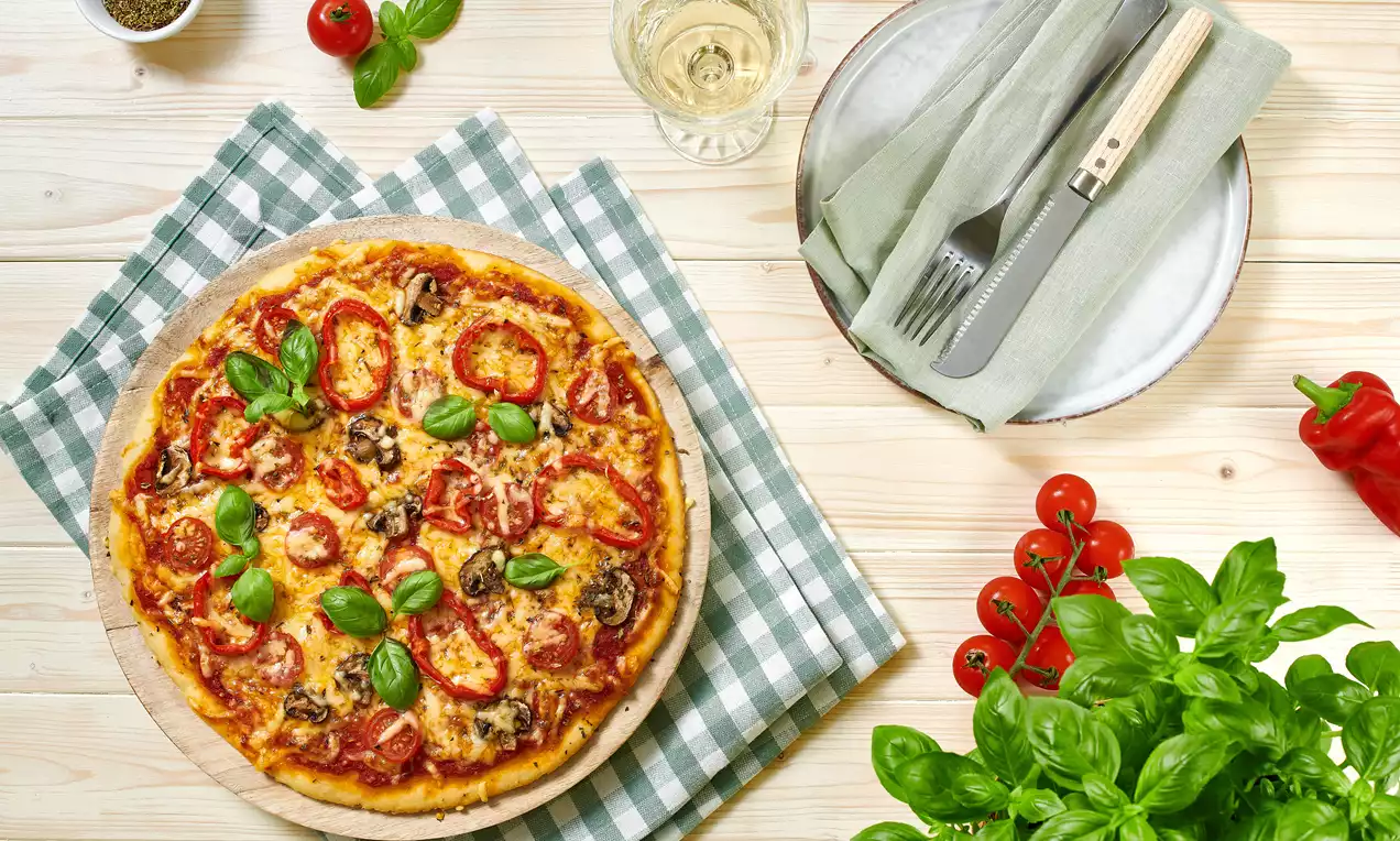 Goldbraune vegetarische Pizza mit Tomaten, Pilzen, Käse und frischem Basilikum auf kariertem Tuch, umgeben von Basilikum und Gemüse