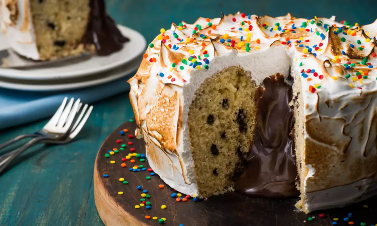 Bolo dourado com massa leve e pontos de gotas de chocolate, coberto com merengue tostado e confeitos coloridos, recheio cremoso escorrendo sobre prato de madeira