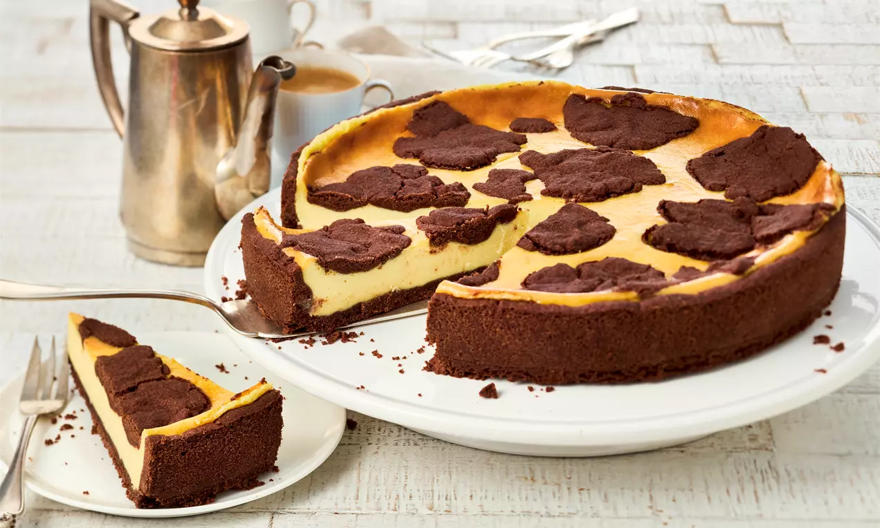 Veganer russischer Zupfkuchen auf weißer Platte, mit heller Quarkfüllung und dunklen Schokostückchen auf der Oberfläche. Ein Stück ist herausgeschnitten und liegt auf einem Teller, daneben Kaffee.