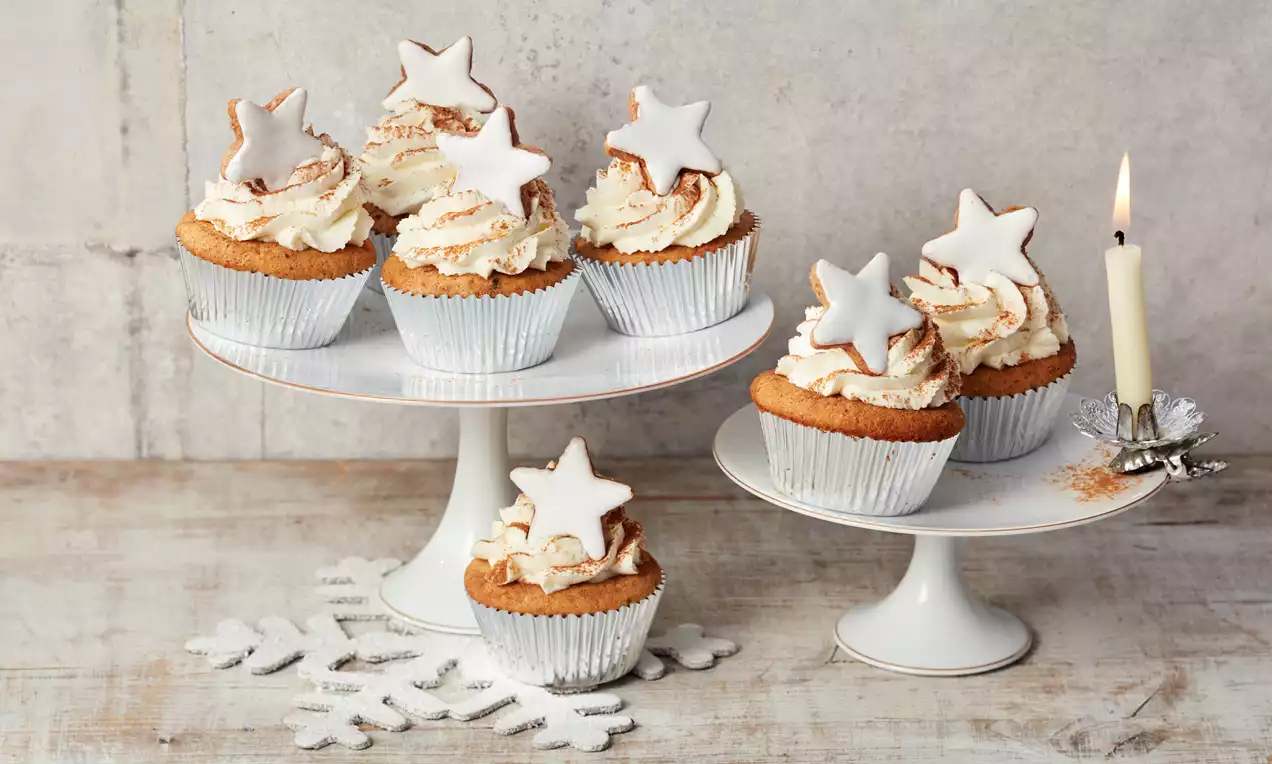 Božićni muffini u bijelim papirnatim košaricama, ukrašeni zavojitim šlagom i bijelim zvjezdicama od glazure na bijelim stalkovima