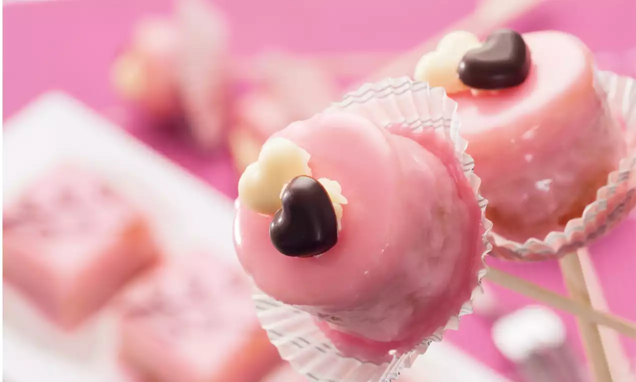 Rosa glasierte Punschkrapfen-Cake-Pops mit glänzender Oberfläche, verziert mit dunklen und weißen Schokoherzen in Papierförmchen auf Holzstielen