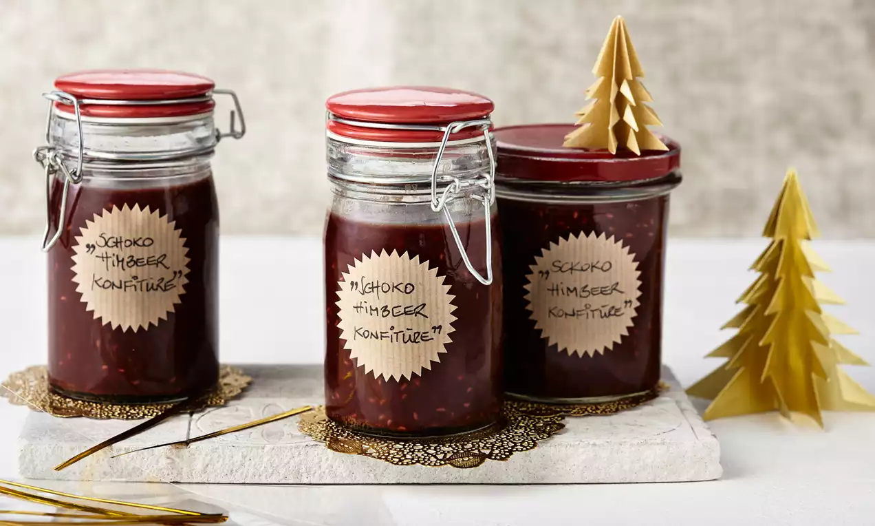 Schokoladige Himbeerkonfitüre im Glas mit leuchtend roten Himbeerstücken, dunkler Schokolade, frischen Beeren und Holzlöffel