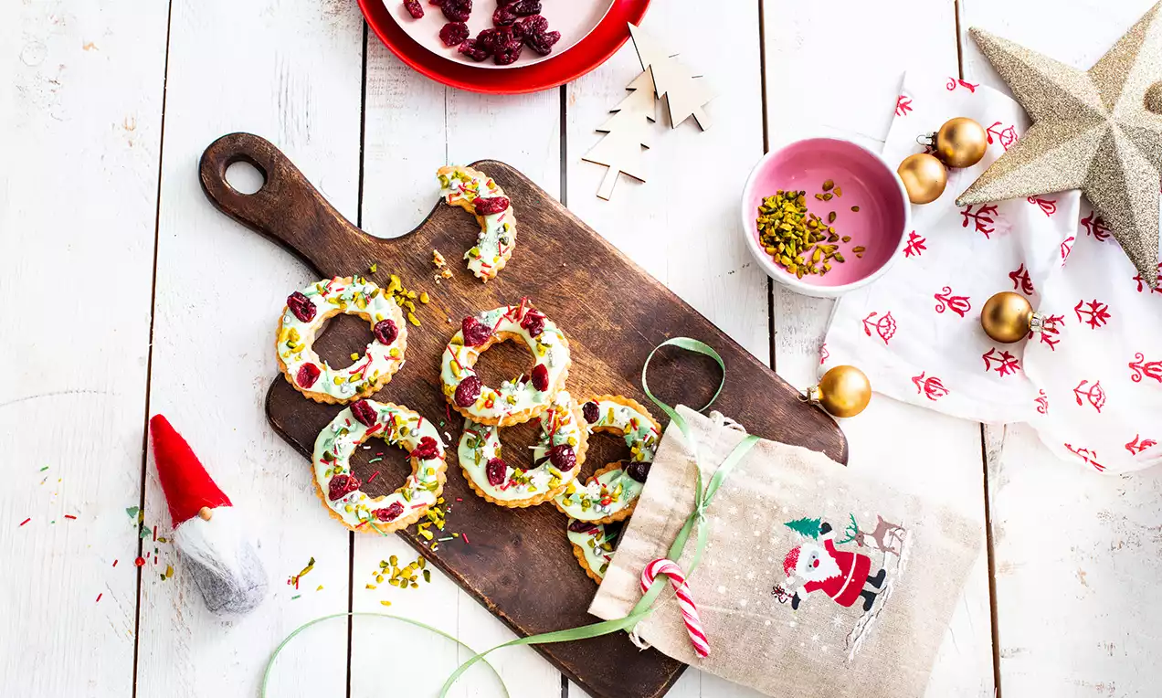 Adventski vjenčići od zlatnosmeđih keksa, premazani bijelom glazurom, ukrašeni sušenim brusnicama i pistacijama na tamnoj drvenoj dasci