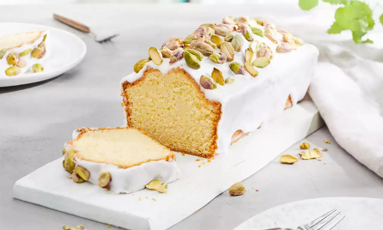 Vanille-Kastenkuchen mit weißem Zuckerguss und grünen Pistazien, angeschnitten auf einem weißen Tablett, im Hintergrund Teller und Besteck