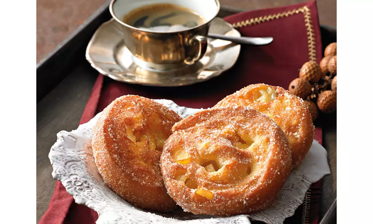 Goldene Apfelberliner mit gezuckerter Oberfläche und leuchtenden Apfelstücken auf zartem Porzellanteller neben einer goldenen Kaffeetasse